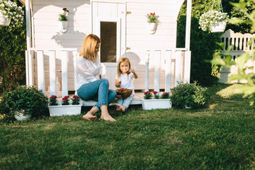 Mother and adorable little kid with bowl of strawberries resting on porch of country house together