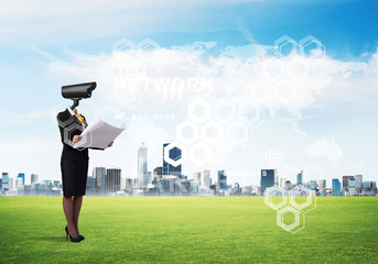 Camera headed woman standing on green grass against modern cityscape