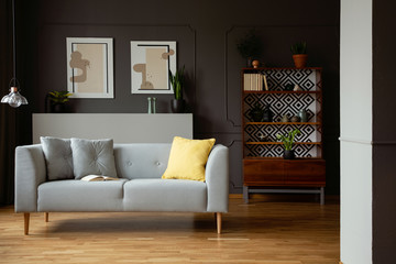 Grey sofa with yellow cushion in vintage living room interior with posters and lamp. Real photo