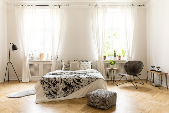 Real photo of a cozy bedroom interior with a double bed, pouf, lamp, windows and chair
