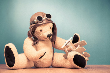 Retro Teddy Bear toy in leather pilot's hat and vintage goggles sitting on the floor and playing with handmade wooden plane front mint green wall background. Old style filtered photo