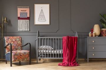 Real photo of a baby crib with a red blanket standing between an arnchair and lamp, and a cupboard with ornaments in child's room interior - obrazy, fototapety, plakaty