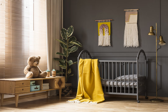 Real photo of a cot with a yellow blanket standing between a low cupboard with a bear and a lamp in baby room interior