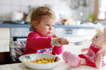 Adorable baby girl eating from fork vegetables and pasta. Little child feeding and playing with toy...
