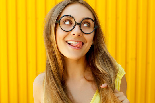 Beautiful young woman in funny toy glasses smiling and showing tongue over yellow background at daylight