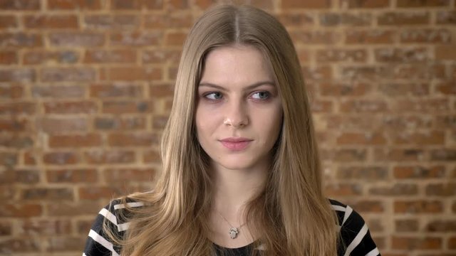 Young cute blonda is watching at camera suspiciously, brick background
