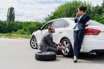 handsome businessman changing tires on car on road, businesswoman talking by smartphone