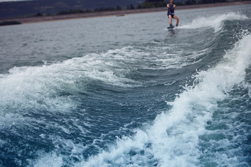 Cropped shot of unrecognizable man riding wakeboard on lake or river behind motor boat. Watersports, vacations, active healthy lifestyle, extreme and adrenaline concept. Selective focus on waves