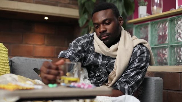 Portrait of likable ill African guy holding cup of tea, drinking. Sick student lying on couch, chatting with friends, smiling. Heat. Illness.