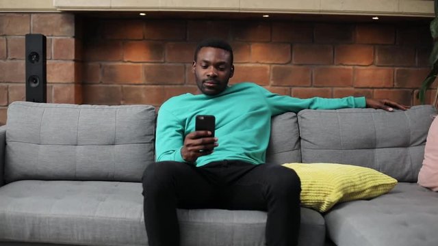 Cute African guy wearing mint sweater sitting on sofa and looking at screen of smartphone. Sick young man having pain in stomach, massaging painful place.
