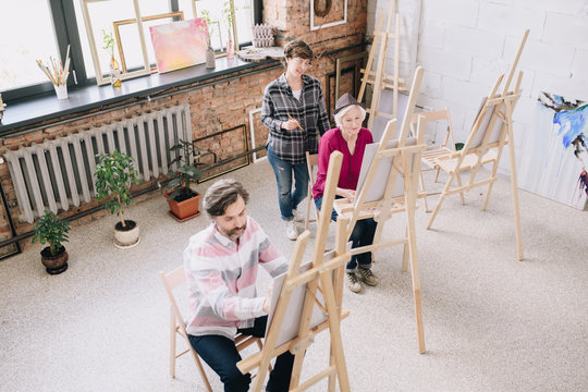 High angle portrait of two adult students  painting sitting by easels in art studio with female art teacher watching them in spacious sunlit loft space, copy space