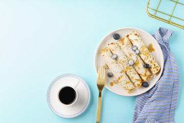 Rolled pancakes with blueberry, caramel and sugar powder. Served with a cup of black coffee on blue background. Copy space