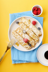 Rolled pancakes with blueberry and raspberry, caramel and powdered sugar on yellow background. Served with black coffee. Top view