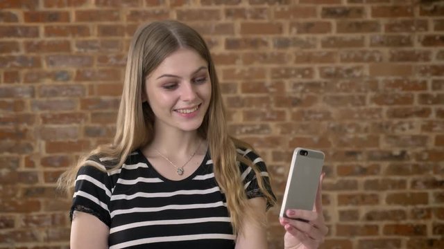 Young happy blonda is talking in video chat on smartphone, waving hello, communication concept, brick background