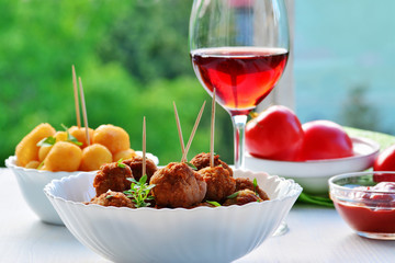 Meatballs in white bowl , glass of red wine cheese balls and tomatoes on white wooden table outdoors.