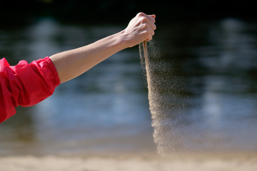 Woman in pink raincoat siting on beach of river and pouring sand, hand close up.