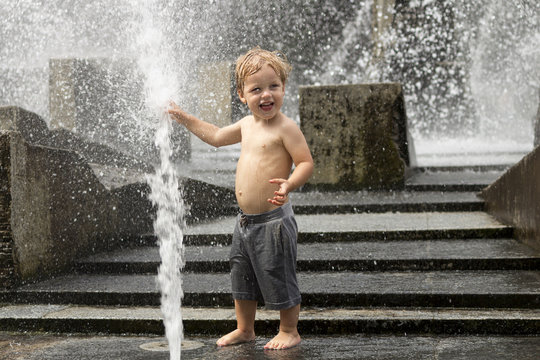 Funny baby boy trying to cauch water stream in fountain. Cute toddler playing in the city fountain.