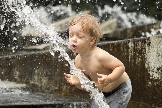 Funny baby boy trying to cauch water stream in fountain. Cute toddler playing in the city fountain.
