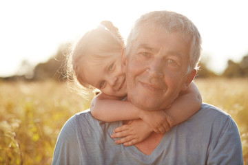 Cute little female kid with gentle smile embraces her grandfather, have joyful expressions, enjoy...