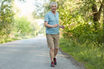 Fototapeta premium Cheerful motivated active male pensioner runs on asphalt, listens music with earphones, enjoys peaceful atmosphere and freah air in countryside, likes sport. People, leisure and fitness concept