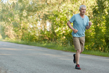 People, retirement and sport motivation concept. Healthy fit grey haired male pensioner runs on asphalt, listens favourite melody in earphones, enjoys leisure time outdoor in rural area. Man runner