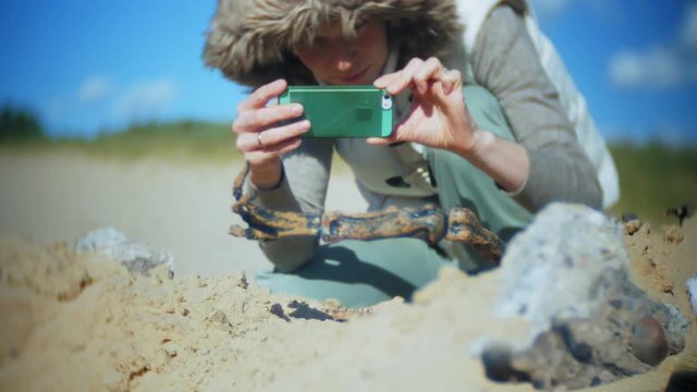 The woman is engaged in excavating bones in the sand, Skeleton and archaeological tools. Makes photo on smartphone