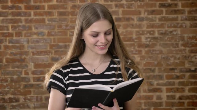 Young sweet blonda is reading book, thumb throudn page, smiling, brick background