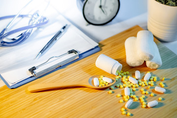 Pills spilling out of pill bottle with gauze bandage roll, pharmaceutical wooden spoon, stethoscope, clipboard and pen on doctor desk. Prescription medicine or medical diagnosis and treatment concepts