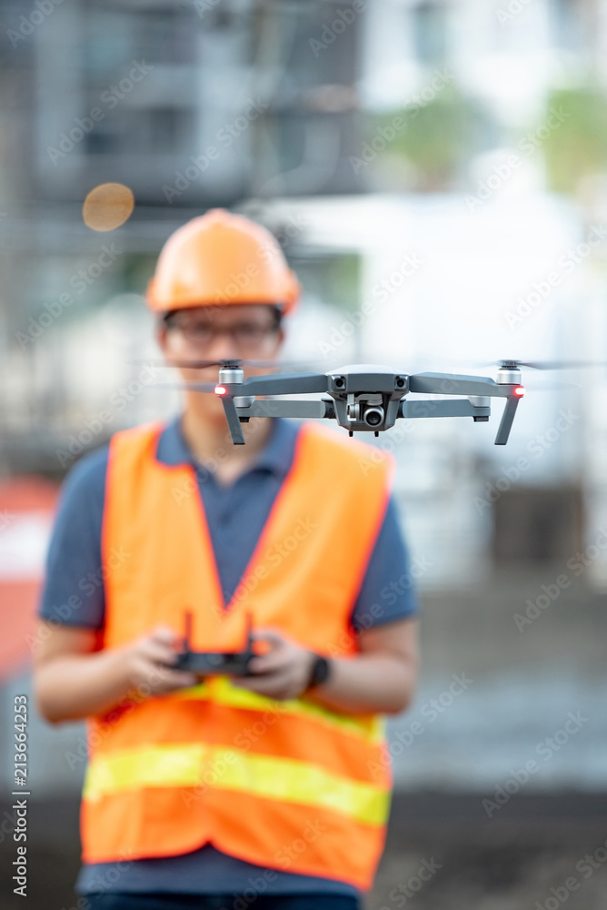 Wall mural young asian engineer man flying drone over construction site. using unmanned aerial vehicle (uav) fo