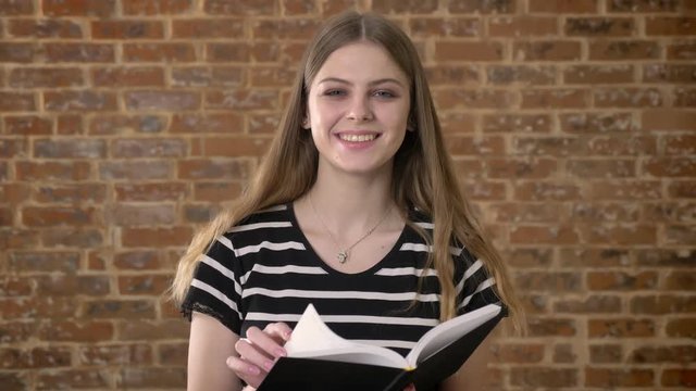 Young beautiful blonda is reading book, watching at camera, smiling, brick background