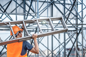 Young Asian maintenance worker with orange safety helmet and vest carrying aluminium step ladder at construction site. Civil engineering, Architecture builder and building service concepts - obrazy, fototapety, plakaty