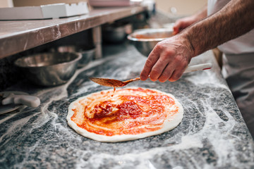 Preparing pizza. Chef's hand adding tomato sauce on pizza dough. - obrazy, fototapety, plakaty