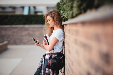 Female student using smart phone and holding books while walking in campus.