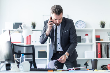 A man is standing near a table in the office, talking on the phone and flipping through documents.