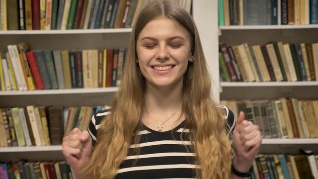Young sweet blonda is watching at camera, smiling, raising hands, library on background
