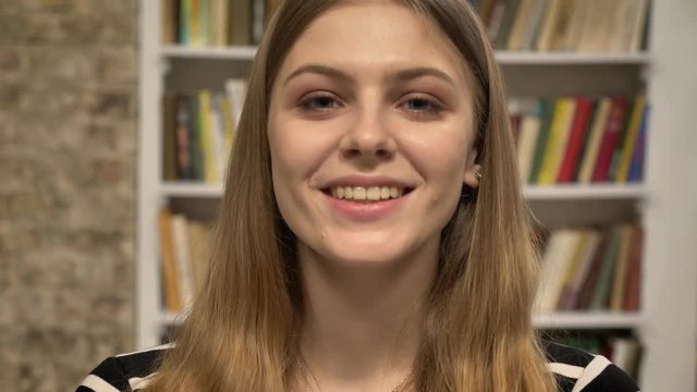 Young sweet blonda is watching at camera, smiling, library on background