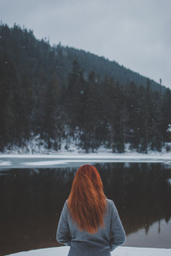 Handsome redhead girl looking in the distance. Splendid shady black mountain lake reflecting trees of forest behind her. Dreamy winter landscape of Carpathian mountains