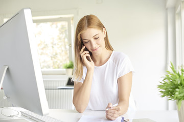 Obraz premium Young professional woman making a call on her mobile phone while working on computer. Home office