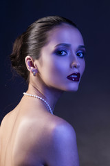 Beautiful brunette girl with naked shoulders and dark red lips make-up, wearing earrings and a pearl necklace, illuminated in blue.