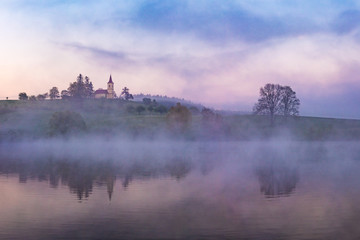 Mystical and dramatic scene. A small on a hill reflected in nearby lake. Beautiful colorful morning light before sunrise. Amazing landscape.