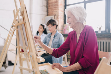 Side view portrait of art students sitting in row and painting at easels in art studio, focus on smiling  senior woman enjoying work copy space