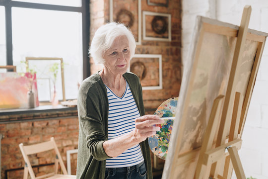 Waist up portrait of white haired senior woman holding palette painting pictures at easel in  art studio standing against windows in sunlight, copy space
