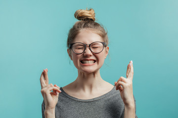 Portrait of overjoyed young caucasian woman with hairbun style lifting fists with crossed fingers from joy or happiness, screwing up her eyes and clenching teeth, celebrating her success