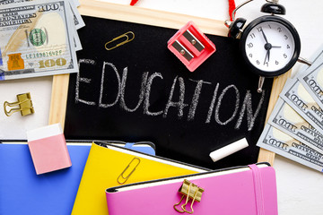 Time & money, word education written on black board, college tuition concept. Pack of new one hundred dollar bills, notebooks, school supplies, alarm clock. Background, copy space, close up, flat lay