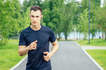 A young handsome man in a black T-shirt runs through the park in his headphones