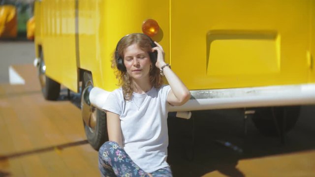 young woman outdoors by yellow vagon car listening music in headphones using smartphone - relaxing, enjoying, concept of technology and travel