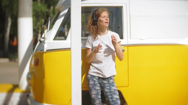 young woman outdoors by yellow vagon car listening music in headphones using smartphone - relaxing, enjoying, concept of technology and travel