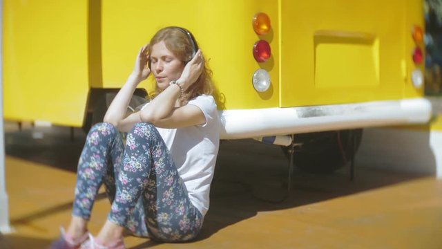 young woman outdoors by yellow vagon car listening music in headphones using smartphone - relaxing, enjoying, concept of technology and travel