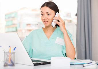 Practicioner woman in medical uniform talking by phone at  laptop