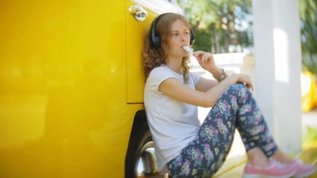 young woman outdoors by yellow vagon car listening music in headphones using smartphone - relaxing, enjoying, concept of technology and travel